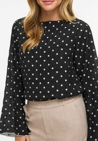 Mustast läbipaistvast kangast pluus valgete täppidega, ruudukujulise kaelusega, pikkade kellukakujuliste varrukatega, tekstuurse kangaga. Sobitatud beežide kohandatud pükstega.