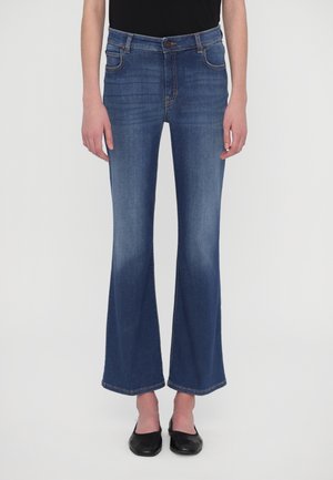 Persoon die blauwe uitlopende spijkerbroeken en zwarte puntige platte schoenen draagt, staand tegen een effen witte achtergrond.