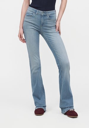 Kvinde iført lyseblå højtaljede flare-jeans og bordeauxsko, står med den ene hånd hvilende på hoften.