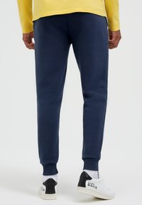 Person trägt marineblaue Jogginghosen, weiße Socken und weiße Turnschuhe mit schwarzen Fersen und "Camp David"-Markierung, steht vor einem einfarbigen Hintergrund.