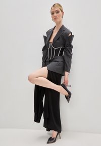 Blazer grigio su misura con dettagli drammatici sulle spalle, corsetto nero, pantaloni neri larghi e décolleté neri scintillanti con dettagli a fiocco.