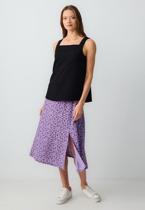 Zwarte mouwloze top met vierkante halslijn, gecombineerd met een lila bloemenrok met een hoge split tot de dij. Witte sneakers maken de look af.