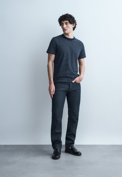 Νεαρός άνδρας με σγουρά μαλλιά που φοράει μαύρο και λευκό ριγέ μπλουζάκι, σκούρα τζιν και μαύρα παπούτσια, στέκεται μπροστά σε ένα απλό ανοιχτόχρωμο τοίχο.