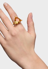 Pierścionek w kolorze złotym z dużym trójkątnym pomarańczowym kamieniem szlachetnym, osadzonym na gładkim pierścieniu, z dwoma małymi okrągłymi akcentami po obu stronach.