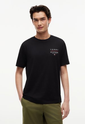 Άνδρας που φοράει μαύρο t-shirt Tommy Hilfiger και ελιά παντελόνι, στέκεται με το ένα χέρι στην τσέπη μπροστά σε ένα απλό φωτεινό φόντο.