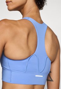 Lys blå sports-BH med racerback-design, preget av dekorativ søm og en kontraststripe i kanten. Glatt, elastisk stoff.