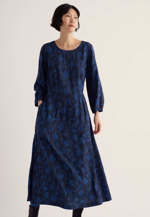 Woman wearing a long dark blue dress with floral pattern and long puffed sleeves, standing against a plain light background.