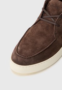 Pantof brown din suede cu vârful rotunjit, având șireturi în ton și o talpă din cauciuc de culoare crem. Prim-plan care arată textură și detalii de cusătură.