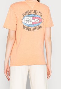 Kortärmad orange T-shirt med "Tommy Jeans Worldwide" grafik i blått och rosa på baksidan; mjukt tyg och avslappnad passform.
