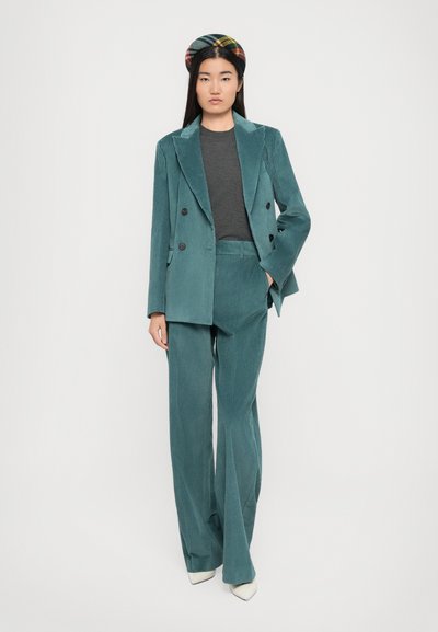 Completo di velluto a coste color teal con giacca doppiopetto e pantaloni a gamba larga, abbinato a un top grigio e a una fascia per capelli a fantasia. Accessori bianchi.
