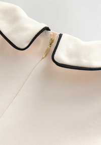 Indumento con colletto in morbido tessuto beige con finitura testurizzata, caratterizzato da un bordo nero e una zip dorata sul retro.