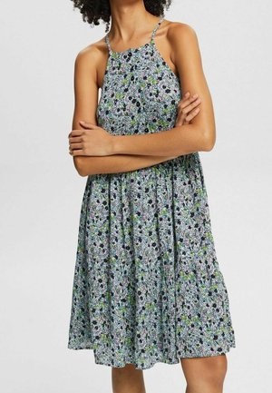Donna che indossa un vestito floreale senza maniche con spalline sottili, braccia incrociate sul petto, in piedi contro uno sfondo chiaro e semplice.