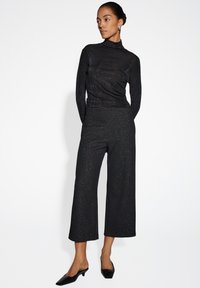 Maglia nera a maniche lunghe con colletto alto, abbinata a pantaloni neri cropped con una leggera brillantezza. Gli accessori includono orecchini grandi e mules neri.