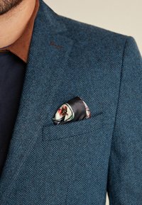Blauer Fischgrätblazer mit braunem Kragen, ausgestattet mit einem gefalteten schwarzen Satin-Einstecktuch mit dekorativem Muster. Strukturierte Stoffdetails sind sichtbar.