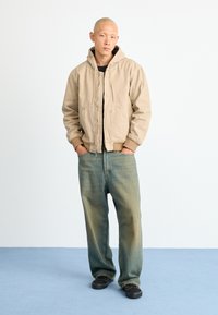 Beige hooded jacket with zipper, black inner layer, light blue jeans, and black shoes. The jeans are relaxed fit with a faded effect.