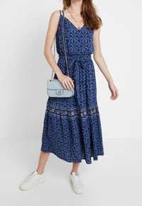Robe maxi bleue à fleurs avec manches courtes, taille cintrée et jupe à volants. Associée à un sac à main bleu clair et des baskets blanches.