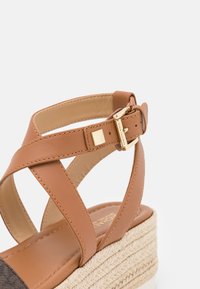 Sandale à plateforme en cuir marron avec des straps croisées, une boucle en métal doré et une semelle enveloppée de jute ; texture lisse et design arrondi.