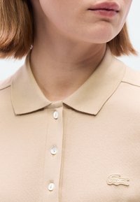 Μπεζ πόλο πουκάμισο με υφασματινή υφή, κλασικό γιακά, τρία κουμπιά και κεντημένο λογότυπο στο αριστερό στήθος.
