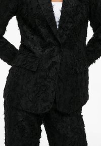 Black suit made of textured fabric with a frayed appearance. Features a single-breasted blazer and straight-leg pants with pockets.