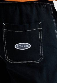 Črne denim hlače z belimi šivi na zadnjem žepu, ki prikazuje sivo ovalno logotip Billabong. Tkanina ima teksturiran končni izgled.