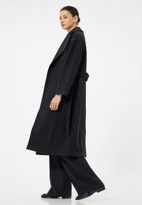 Cappotto nero in tessuto testurizzato, con colletto a scialle, maniche ampie, cintura da annodare e tasche laterali, indossato su pantaloni neri a gamba larga.