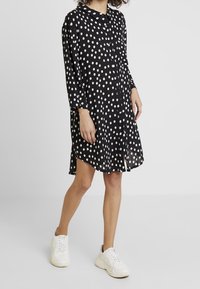 Schwarzes Hemdkleid mit weißen Polka-Dots, Knopfleiste vorne, lange Ärmel, abgerundeter Saum, kombiniert mit weißen klobigen Sneakern.