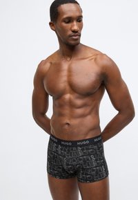 Μαύρα ανδρικά εσώρουχα boxer με επαναλαμβανόμενο μοτίβο λογότυπου, ελαστική μέση με τη σήμανση "HUGO", λεία υφή. Το μοντέλο ποζάρει με τα χέρια πίσω από την πλάτη.