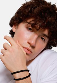 Black braided leather bracelets on a wrist. The individual has tousled brown hair and is wearing a plain white shirt, with a neutral expression.