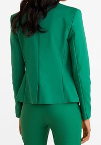 Blazer verde su misura realizzato in tessuto morbido, caratterizzato da un design aderente, retro peplum e rever a punta. Abbinato a pantaloni coordinati.