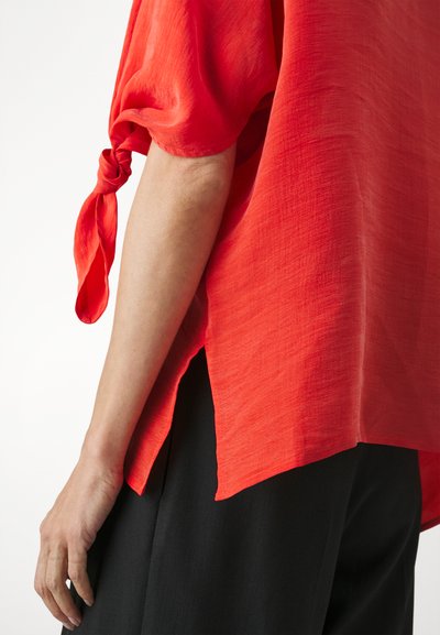 Red, textured short-sleeve top with a tied cuff and side slit, paired with black fabric pants. Simple, flowing design.