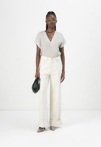Lichtgrijze v-hals blouse, brede crèmekleurige broek met opgerolde manchetten, zwarte handtas en taupe hakken. Glad materiaal met een gestructureerde pasvorm.
