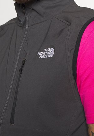 Grå vest med lynlåslukning foran, med The North Face-logo i hvid. Kontrastfulde sorte armbånd, båret over en lyserød t-shirt med korte ærmer.