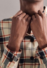 Camicia di cotone a quadri con colletto button-up; presenta motivi rossi, navy e beige; la texture è liscia con bottoni neri a contrasto.