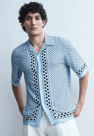 Jeune homme aux cheveux bouclés portant une chemise à boutons bleu clair avec des motifs de cœurs noirs, debout les mains dans les poches devant un fond uni.