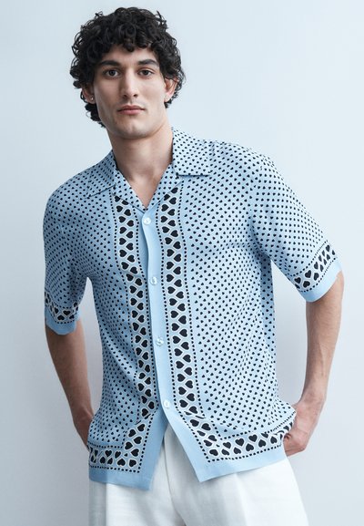 Jeune homme aux cheveux bouclés portant une chemise à boutons bleu clair avec des motifs de cœurs noirs, debout les mains dans les poches devant un fond uni.