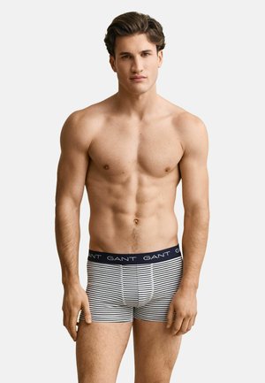 Homme aux cheveux courts et foncés portant un boxer rayé bleu marine et blanc avec le logo "GANT" sur la ceinture, debout devant un fond uni.