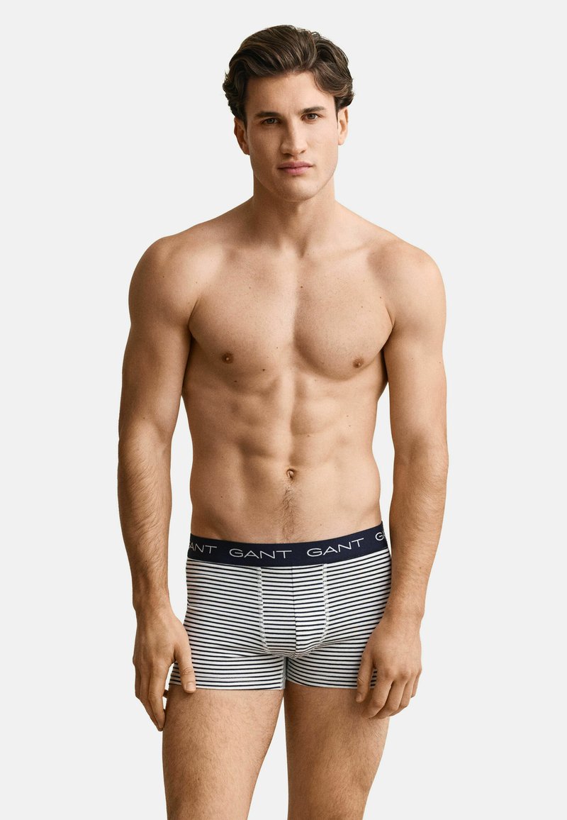 Homme aux cheveux courts et foncés portant un boxer rayé bleu marine et blanc avec le logo "GANT" sur la ceinture, debout devant un fond uni.