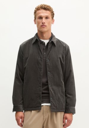 Junger Mann mit lockigem Haar, der eine dunkelgraue Cordjacke über einem dunklen Poloshirt und beigen Hosen trägt, steht vor einem einfarbigen Hintergrund.