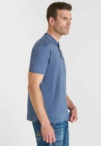 Polo shirt blu con colletto, maniche corte e strisce bianche di accento. Indossato con jeans blu, che mostrano una vestibilità pulita e sartoriale.