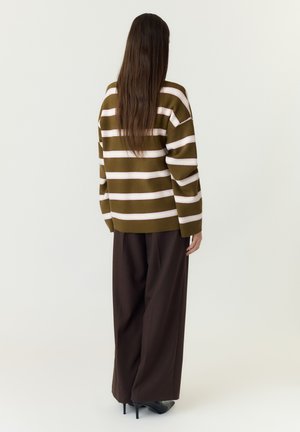 Femme aux longs cheveux bruns portant un pull rayé olive et blanc, un pantalon large marron et des chaussures à talons noirs, de dos.