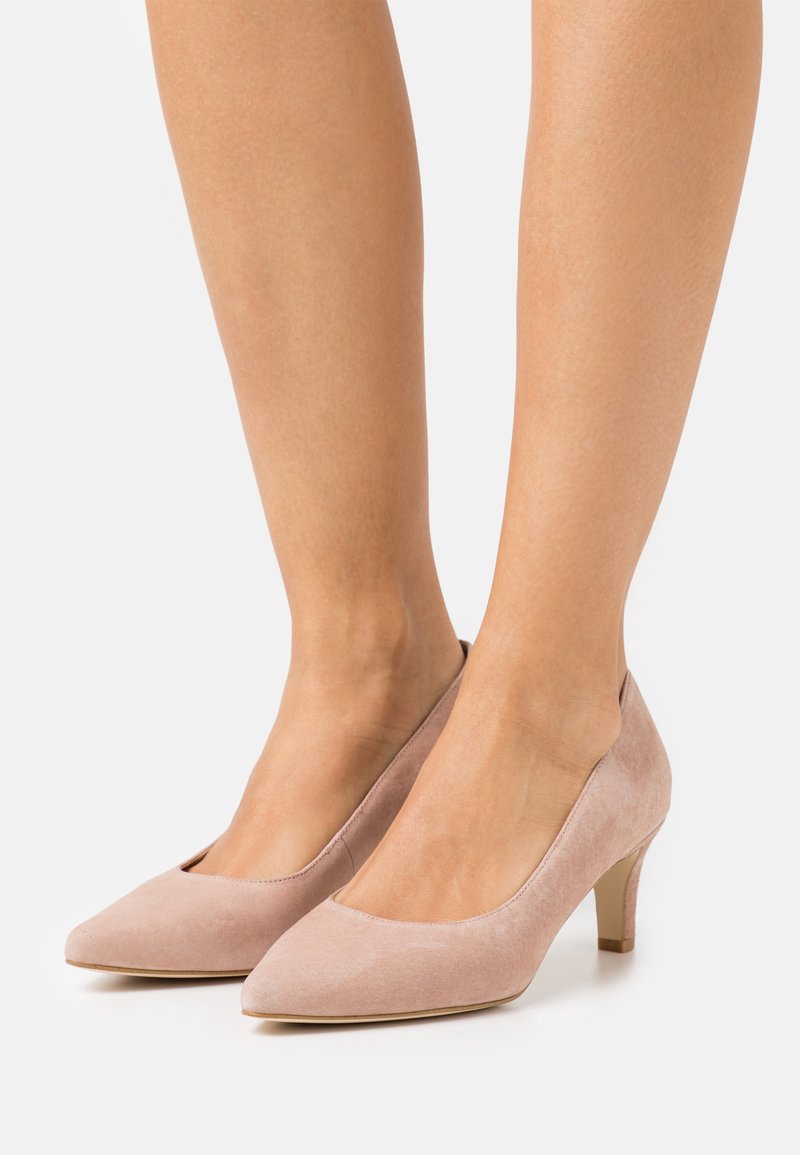 Jambes portant des escarpins en daim rose clair à petit talon avec bouts pointus sur un fond uni.