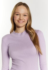 Helllila, gerippte Langarmbluse mit hohem Kragen und glatter Textur, die eine figurbetonte Silhouette aufweist. Haare in lockeren Wellen gestylt.