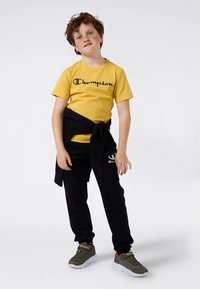 Chlapec s kudrnatými vlasy, oblečený v žlutém tričku Champion, černých teplákách, zelených sneakers a s černou mikinou uvázanou kolem pasu.