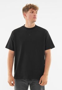 Černé bavlněné tričko s kulatým výstřihem, krátkými rukávy a diskrétním logem embosovaným uprostřed. Standardní střih s hladkou texturou.