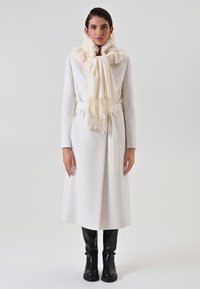 Manteau blanc en mélange de laine avec une taille ceinturée, associé à une écharpe crème avec un col en fourrure et de longues franges. Bottes noires montantes.