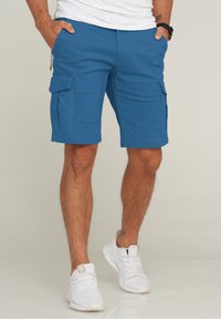 Blaue Cargo-Shorts aus Stoff, mit Seiten Taschen und knielanger Passform. Kombiniert mit weißen Sportschuhen.