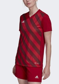 Rotes Sporttrikot mit dunkelroten diagonalen Streifen, kurzen Ärmeln und V-Ausschnitt. Kombiniert mit passender roten Shorts, die ein Adidas-Logo tragen.
