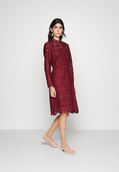 Rochie din dantelă burgundă cu mâneci lungi, având motive florale complexe și un tiv ondulat. Asortată cu sandale cu toc alb și curea.