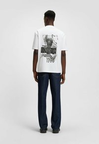 Man wearing white graphic t-shirt with black text and statue image, dark blue jeans, and black shoes, standing against plain background.