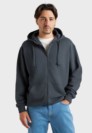 Homme aux cheveux foncés et boucles d’oreilles portant un sweat à capuche gris foncé zippé sur une chemise blanche et un jean bleu clair, posant avec une main dans la poche.
